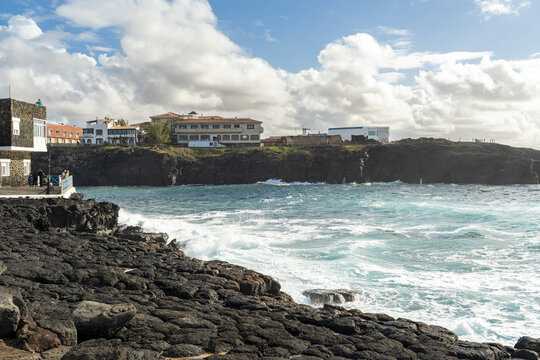 View of the rugged volcanic coastline and turquoise waves crashing against black rocks with white buildings perched on the cliffside in El Cotillo, Canary Islands, Spain.