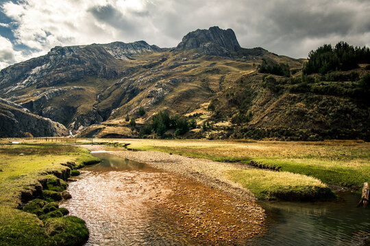 View of Huascaran mountain rising above a winding river and grassy plains under a dramatic cloudy sky in Yungay Province, Ancash, Peru.