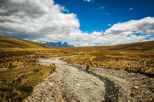 View of the rocky stream bed winding through the dry Andean plateau with the snow-capped Huascaran mountain in the distance under a bright blue sky in Yungay Province, Ancash, Peru.