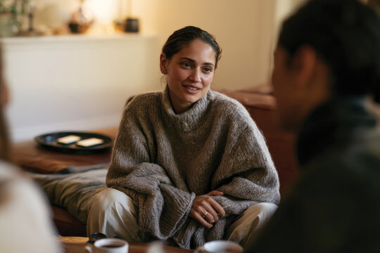 A young woman in a warm oversized sweater smiles during a relaxed gathering, epitomizing comfort, connection, and warmth among friends in a cozy environment.