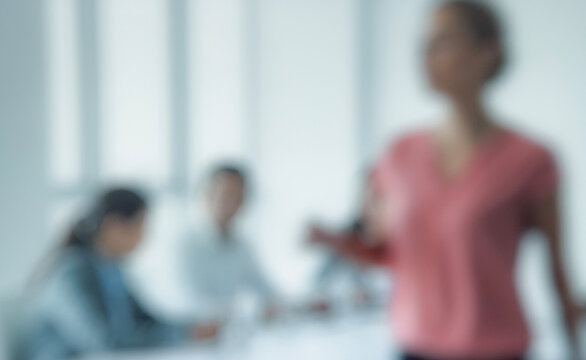 A blurred image of a professional meeting in a bright office, featuring a focused individual speaking while others engage in discussion in the background.