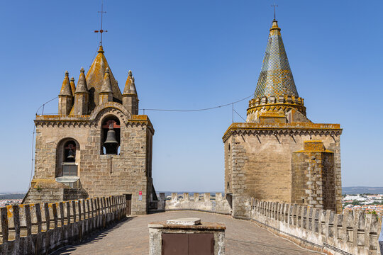 View of the Cathedral of Evora with its stone towers, bell tower, and tiled spire rising above the historic battlements under a clear blue sky in Evora, Distrito de Evora, Portugal.