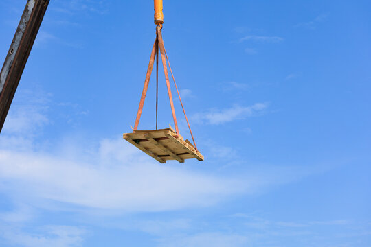 A construction crane lifts a wooden pallet secured with straps against a clear blue sky, illustrating industrial logistics, cargo transport, and heavy lifting at a building site.