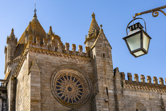 View of the Cathedral of Evora with its intricate rose window, stone walls, and crenellated roof under a clear blue sky, featuring a hanging lantern in Evora, Distrito de Evora, Portugal.