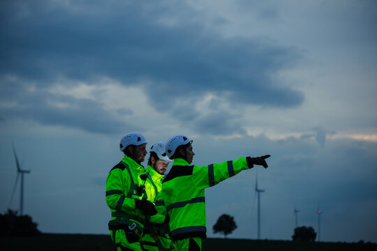 Silhouetted wind energy technicians in high visibility jackets stand under a dramatic dusk sky near wind turbines, one pointing ahead, symbolizing teamwork in renewable energy work.
