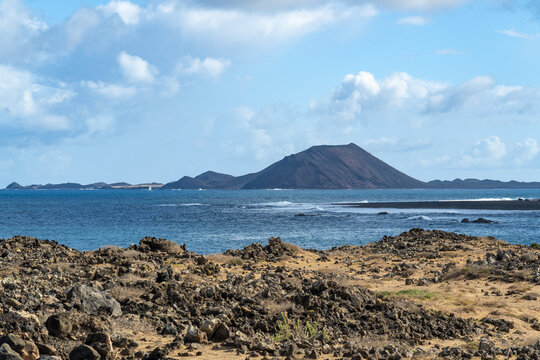 View of the volcanic landscape of Isla de Lobos across the blue sea from the rocky coast of Corralejo, Canary Islands, Spain.