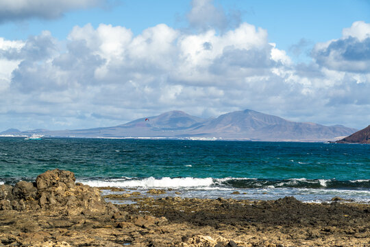 View of the rocky coastline and turquoise sea with volcanic mountains in the distance under a cloudy blue sky in Corralejo, Canary Islands, Spain.