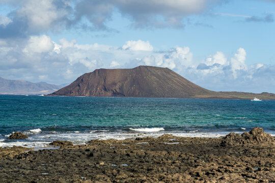 View of the volcanic landscape of Isla de Lobos rising from the turquoise sea with rugged volcanic rocks in the foreground under a blue sky in Corralejo, Canary Islands, Spain.
