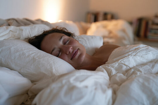 A woman lies in bed with a serene expression, enveloped in soft bedding, evoking feelings of comfort, tranquility, and inner peace in a calming environment.
