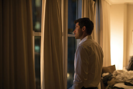 A well-dressed man in a white shirt gazes thoughtfully out the window, embodying contemplation and reflection amidst the comfort of a stylish room.