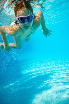 Girl in purple goggles swimming directly toward camera underwater. Concept of swim schools, kids sports and aquatic products.