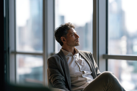 A man thoughtfully gazes out the window in a high-rise office setting, embodying moments of inner reflection amidst a bustling urban landscape filled with possibilities.