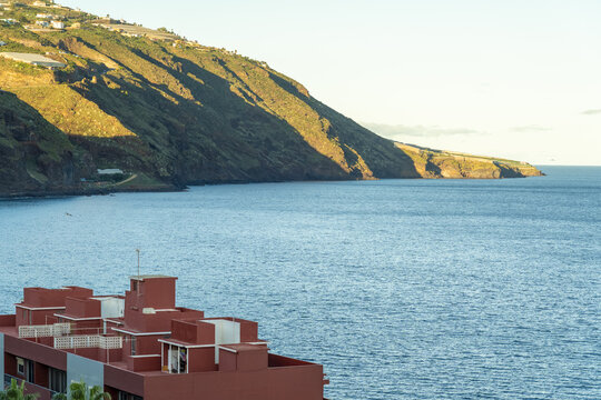 View of a modern red building overlooking the deep blue Atlantic Ocean with a steep mountain slope in the background in Santa Cruz de La Palma, Canary Islands, Spain.