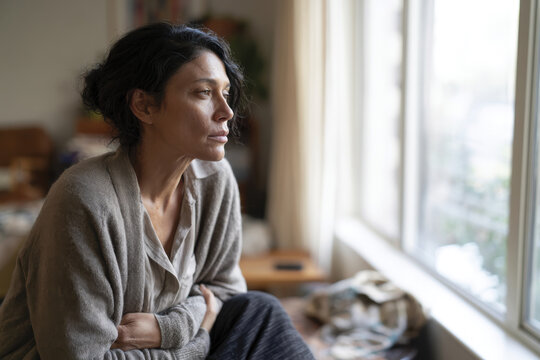 A woman sits pensively in a sunlit room, wrapped in a warm cardigan, reflecting quietly by the window, creating an atmosphere of calm and introspection.
