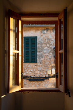 View of a rustic stone wall and a green shuttered window seen through an open wooden window frame in the historic village of Fornalutx, Balearic Islands, Spain.