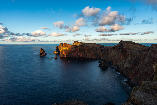 Stunning aerial view of rugged volcanic cliffs and ocean at Ponta de Sao Lourenco, Madeira island, illuminated by warm golden sunset light under a scenic blue sky.