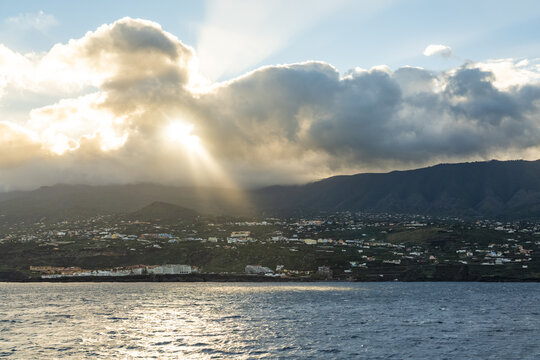 View of sun rays breaking through clouds over the coastal town with mountains in the background and reflections on the sea, Santa Cruz de La Palma, Canary Islands, Spain.