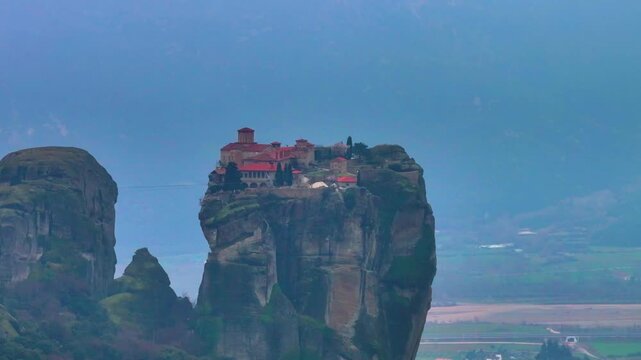 Telephoto shot of the Holy Trinity Eastern Orthodox Monastery built atop a massive rock pillar with Thessaly valley in the background under soft atmospheric light in Meteora, Thessaly, Greece.