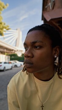 Young African American man sitting outdoors on city street, looking away with bridge in background. Footage is suitable for youth lifestyle ads, urban fashion campaigns, or social media, vertical