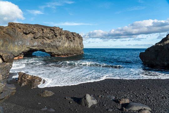 View of a natural volcanic rock arch over a black sand beach with turquoise ocean waves and a clear blue sky in Santa Cruz de Tenerife, Canary Islands, Spain.