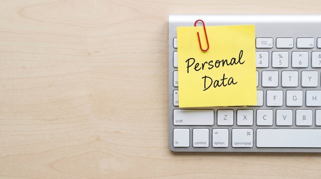 A bright yellow sticky note with a reminder about personal data security and privacy, placed on a modern computer keyboard on a wooden desk
