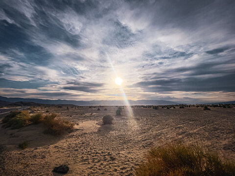 View of Death Valley desert landscape with sparse shrubs, sandy terrain, and a bright sun shining through dramatic clouds in Inyo County, California, United States.