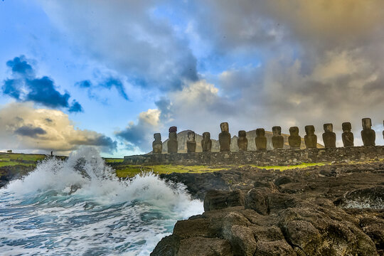 View of the iconic Moai statues standing on the rocky coastline of Easter Island with waves crashing against the shore under a dramatic cloudy sky in Easter Island, Valparaiso, Chile.