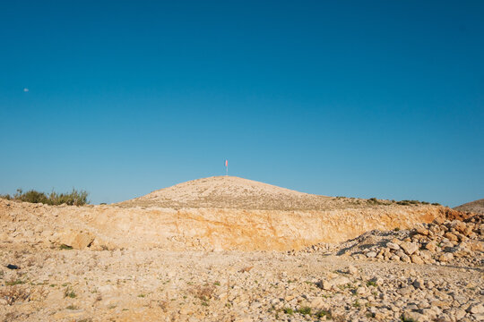 View of a rocky hill with a small flag on the summit under a clear blue sky near the Misnjak Ferry Port in Barbat na Rabu, Primorsko-goranska zupanija, Croatia.