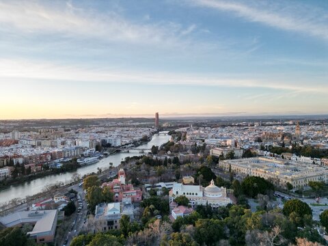 Drone aerial view of Seville, Spain, showing the Guadalquivir River, Torre Sevilla skyscraper, Mar&iacute;a Luisa Park, and historic architecture at sunset