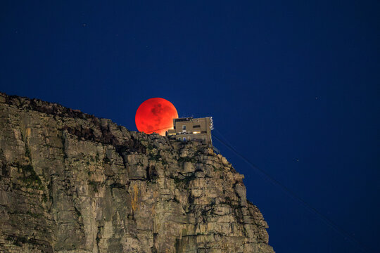 Cape Town, South Africa - 28 August 2026: View of Table Mountain with a blood moon rising behind the cableway station on the rocky cliff edge under a dark blue night sky.