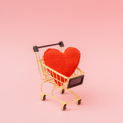 Miniature shopping cart holding a red fabric heart against a pastel pink studio backdrop, photographed from a front three-quarter view. Romantic purchase concept with soft minimal styling. © Bojan