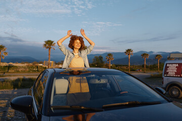 Fototapeta premium A young woman with curly hair stands through a sunroof, arms raised, in a bright desert scene with palm trees, conveying freedom and carefree mood.