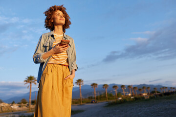 Naklejka premium Smiling woman outdoors in casual summer outfit, striped top, denim jacket, and mustard skirt. Palm trees line a desert backdrop under a clear blue sky. Lifestyle portrait emphasizing confidence,