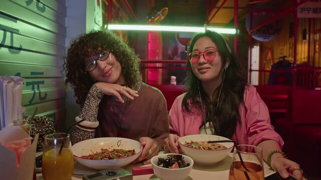 Portrait of two young adult Asian women sitting in Asian restaurant, smiling at camera while enjoying noodles. Clip is perfect for multicultural lifestyle and food delivery marketing campaigns