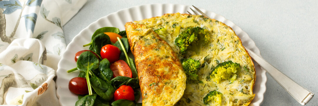 A potato omelette made with eggs and mixed greens is served on a white plate. Spinach, cherry tomatoes, and broccoli are arranged on the side for a healthy breakfast option, banner