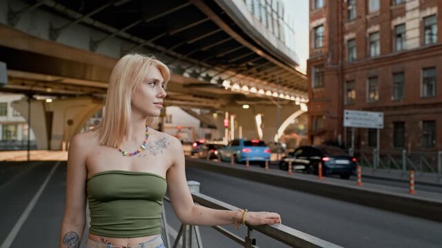 Young adult Caucasian woman standing near metal railing under city bridge, looking away and contemplating. Footage is suitable for urban lifestyle, youth culture, and modern city living promos