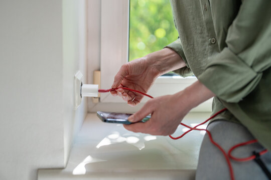 Woman bends damaged USB cable beside charger, searching for working angle to restore unstable smartphone charging while financial hardship delays replacement and forces use of worn accessory at home.