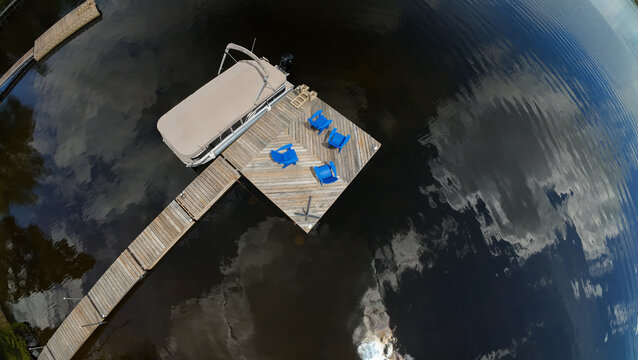A high angle overhead drone view tracks over a private wooden dock holding blue adirondack chairs and a covered pontoon boat on a calm dark lake surface on a beautiful day.
