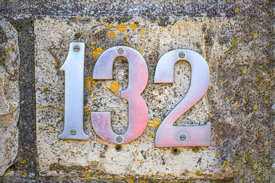 House number 132 sign made of metal digits screwed onto a lichen speckled stone wall, showing one three two numerals in polished address plate design