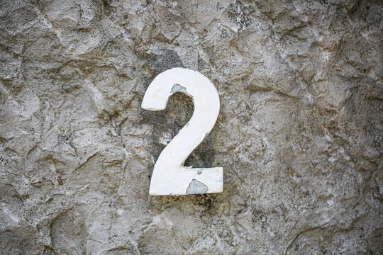 House number two sign in weathered metal, fixed on a rough concrete wall, showing a single peeling painted numeral with rustic aged texture outdoors