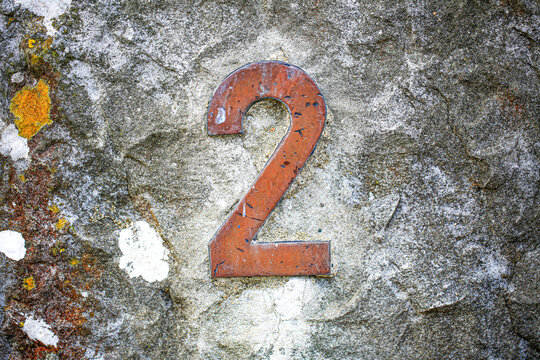 House number two sign in weathered metal, fixed on a rough concrete wall, showing a single peeling painted numeral with rustic aged texture outdoors