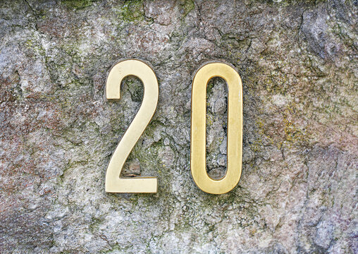 House number 20 sign in shiny brass digits, mounted on a rugged mossy stone wall, showing two and zero numerals with polished metallic address plate finish