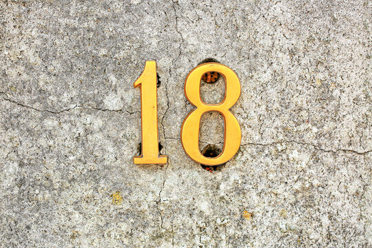 House number 18 sign in polished brass digits, mounted on a cracked concrete wall, displaying one and eight numerals with warm metallic address plate finish