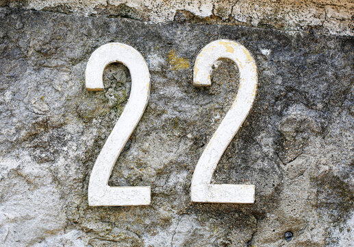 House number 22 sign in peeling painted metal digits, fixed on a rough stone wall, displaying two identical numerals with vintage weathered rustic finish