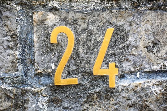 House number 24 sign in shiny gold digits, mounted on a rugged stone wall, displaying two and four numerals with polished metallic address plate design
