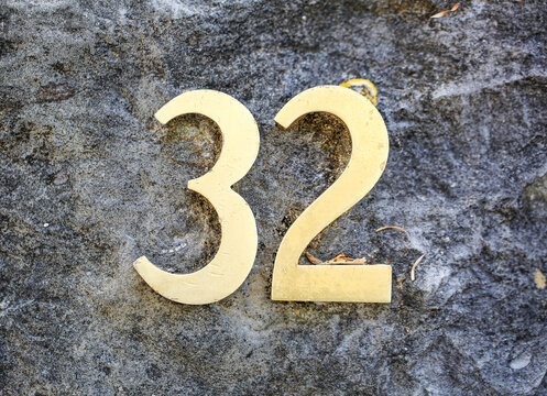 House number 32 sign in shiny gold digits, resting on a rugged concrete surface, showing three and two numerals with polished metallic address plate finish