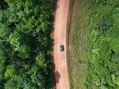Aerial drone view of a car traveling along a red dirt road surrounded by lush tropical vegetation in French Guiana.
