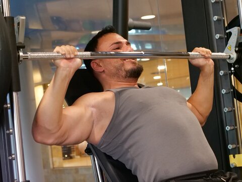 Muscular man in grey tank top performing a seated chest press exercise on a Smith machine in a gym, pushing a barbell upwards.