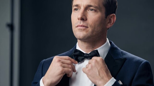 Elegant man in a tuxedo adjusting his black bow tie in a dark studio. Confident businessman in a formal suit getting ready for an event. Luxury style, wealth and success