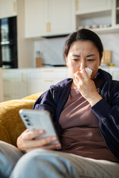 Ill woman blowing nose while using smartphone at home